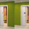 Two ECLISSE Self-Closing Systems for Pocket Doors, one opaque with an orange arrow and one transparent with a blue arrow, are set in a green wall and partially open to reveal a colourful room with curved furniture and shapes inside.