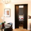 Modern living room with a round black dining table, four chairs, potted orchid, chandelier, and an ECLISSE Pocket Door System - Classic Double Wiring-Ready revealing a cosy bedroom with patterned bedding and red accent cushions.