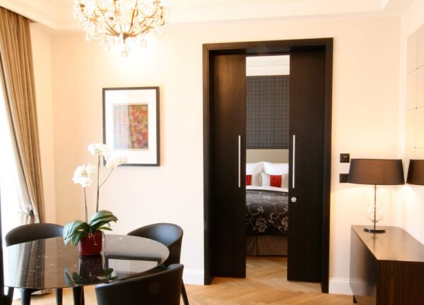 Modern living room with a round black dining table, four chairs, potted orchid, chandelier, and an ECLISSE Pocket Door System - Classic Double Wiring-Ready revealing a cosy bedroom with patterned bedding and red accent cushions.