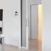 A modern white room features the ECLISSE Pocket Door System - Classic Single Wiring-Ready, partially open to a softly lit corridor. Nearby, a black-and-white portrait print stands beside a small table and white chair.