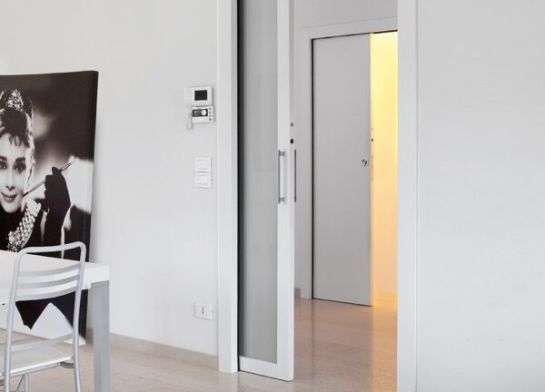 A modern white room features the ECLISSE Pocket Door System - Classic Single Wiring-Ready, partially open to a softly lit corridor. Nearby, a black-and-white portrait print stands beside a small table and white chair.