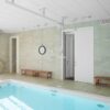 Indoor swimming pool with clear water, two wooden benches along tiled walls, a clock, flotation rings, and an open Classic Hoist Pocket Door System leading to another room. Ceiling rails and ample lighting are also visible.