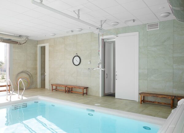 Indoor swimming pool with clear water, two wooden benches along tiled walls, a clock, flotation rings, and an open Classic Hoist Pocket Door System leading to another room. Ceiling rails and ample lighting are also visible.
