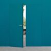 A teal ECLISSE Door System - Syntesis® Hinged Double, nearly matching the wall, stands slightly ajar, revealing an outdoor view of trees and a patio through the opening.
