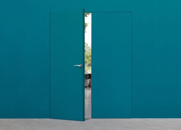 A teal ECLISSE Door System - Syntesis® Hinged Double, nearly matching the wall, stands slightly ajar, revealing an outdoor view of trees and a patio through the opening.