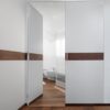 A modern bathroom with an ECLISSE Door System - Syntesis® Hinged Double featuring a horizontal dark wood stripe matching the wooden floor. A bidet and toilet are visible, lit by bright natural light from a window.