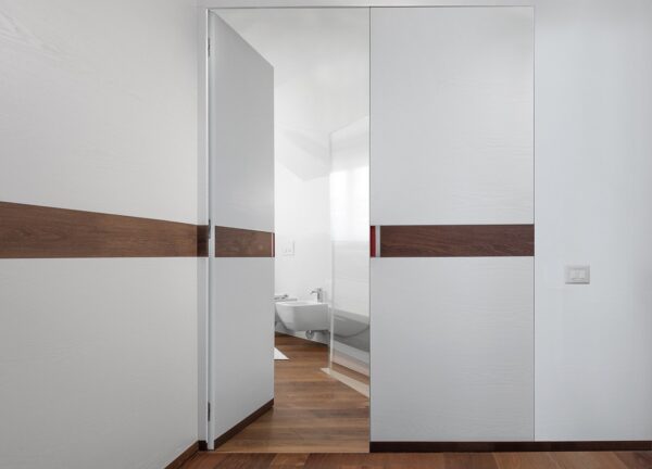 A modern bathroom with an ECLISSE Door System - Syntesis® Hinged Double featuring a horizontal dark wood stripe matching the wooden floor. A bidet and toilet are visible, lit by bright natural light from a window.