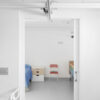 A minimal hospital room with white walls, a single bed with a blue blanket, a small bedside table, and a red chair is seen through a Classic Hoist Pocket Door System, with part of a shower visible on the right.
