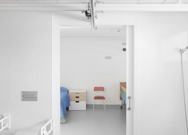 A minimal hospital room with white walls, a single bed with a blue blanket, a small bedside table, and a red chair is seen through a Classic Hoist Pocket Door System, with part of a shower visible on the right.