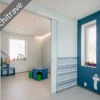 A modern playroom with blue and white walls, playful creature decals, toy shelves, and a window. Features the ECLISSE Pocket Door System - Syntesis® Double with no visible architrave. Text: No architrave.