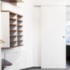 A modern, minimal wardrobe with open shelves, hangers, a dressing gown, and an ECLISSE Wall Mounted Sliding Door Kit and Pelmet–2m Track–Paris 2 partially revealing a bright bathroom with bath and window. Clean white walls complete the space.