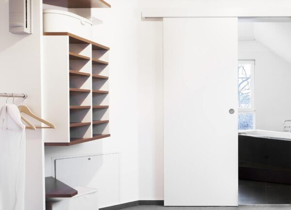 A modern, minimal wardrobe with open shelves, hangers, a dressing gown, and an ECLISSE Wall Mounted Sliding Door Kit and Pelmet–2m Track–Paris 2 partially revealing a bright bathroom with bath and window. Clean white walls complete the space.