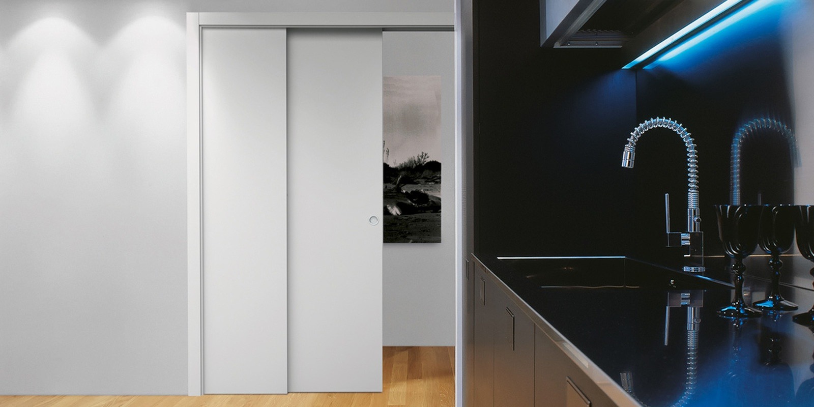 A modern kitchen with black worktops, a flexible tap, blue under-cabinet lighting, and three black goblets. In the background are sleek ECLISSE sliding doors and a partial view of a hallway with wooden flooring.