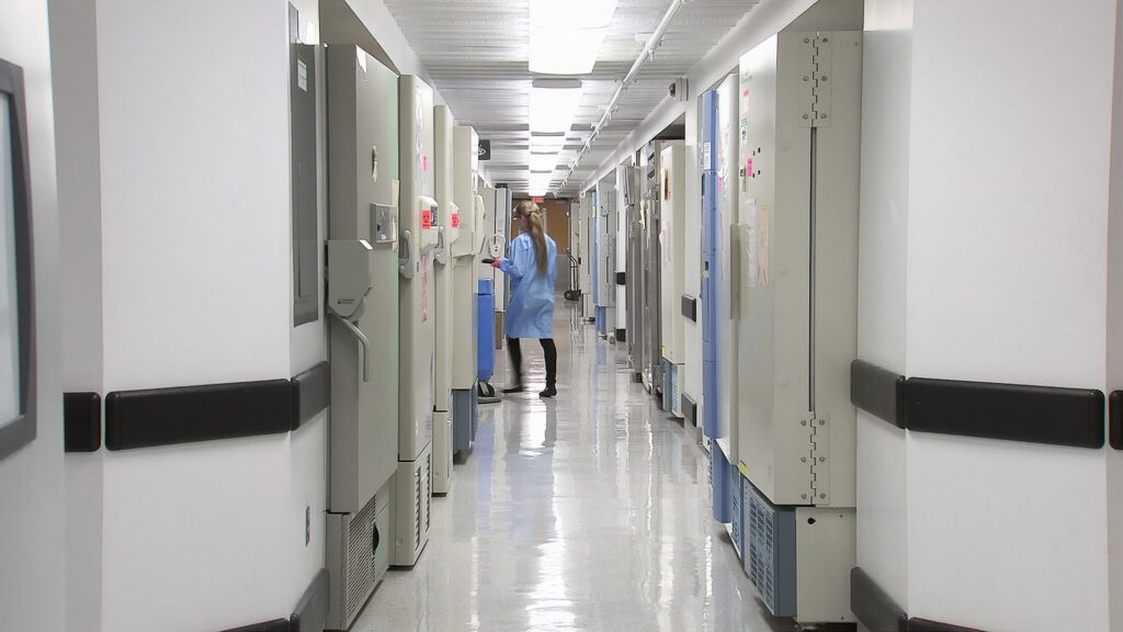 A person in a lab coat stands in a bright, clean corridor with large scientific freezers on both sides and sleek ECLISSE sliding doors leading to different sections of the laboratory or research facility.