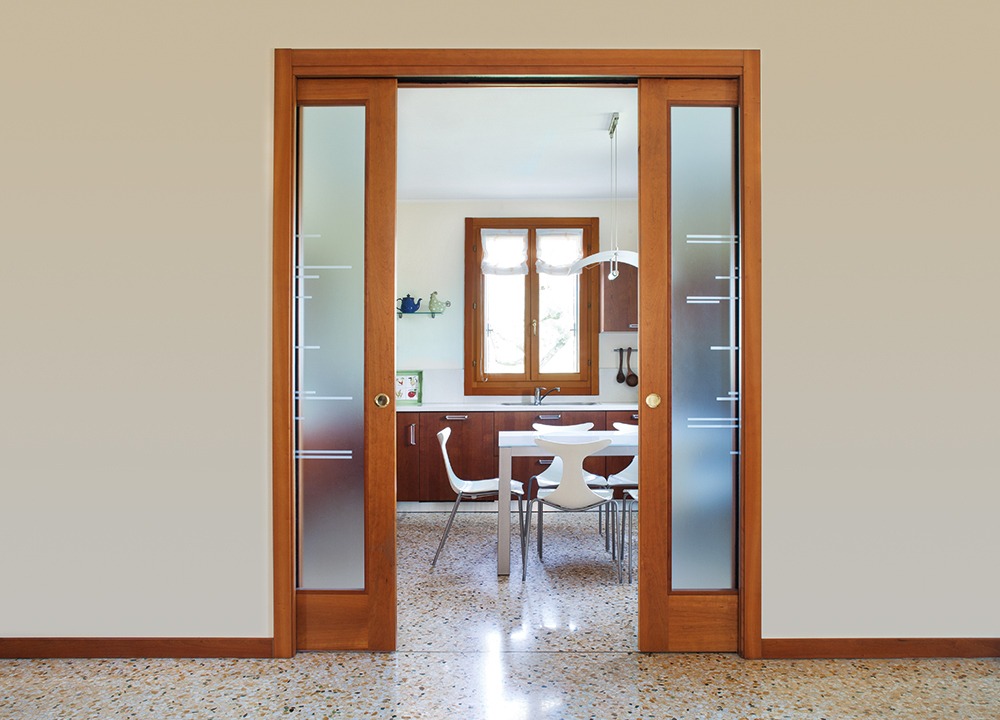 Modern wooden sliding doors with frosted glass panels, like those from ECLISSE, open to reveal a bright kitchen with a white table, white chairs, wooden cupboards, and a window letting in natural light. The floor is terrazzo.