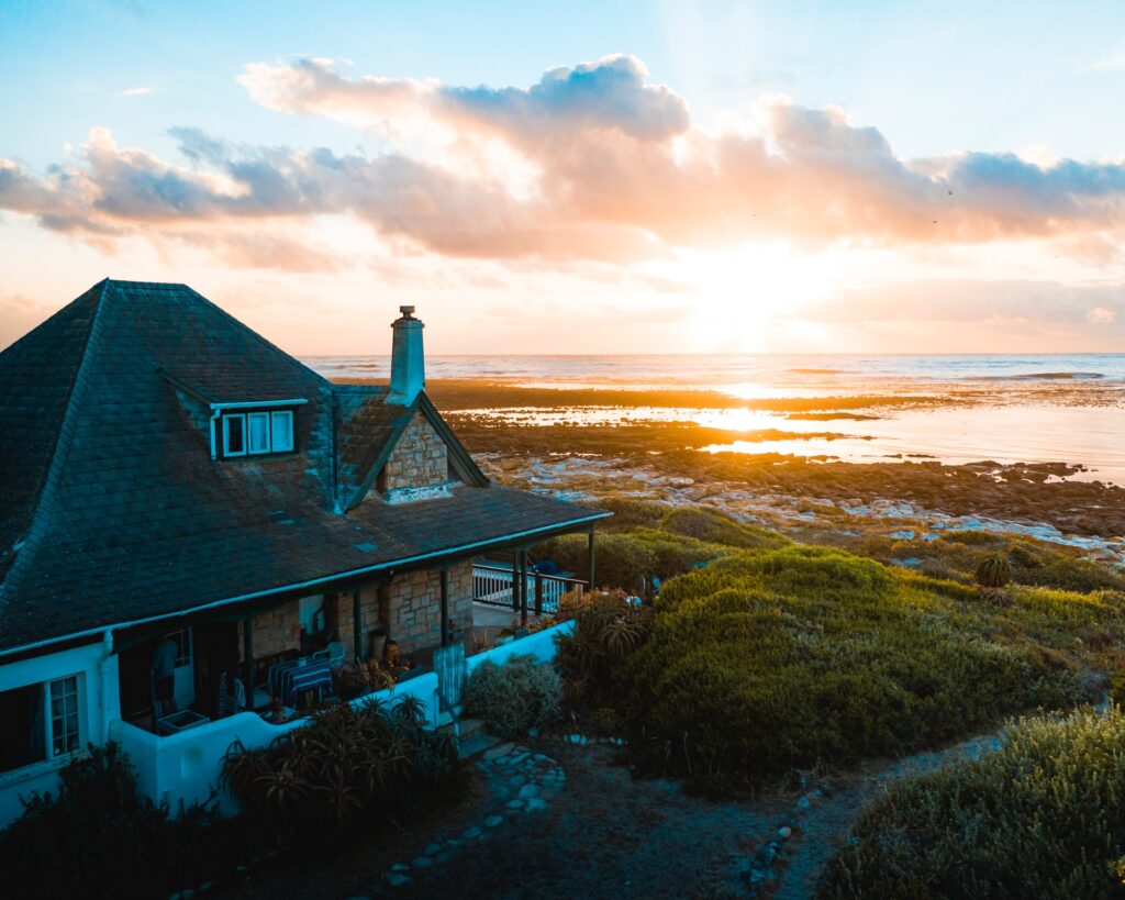 A cosy house with a stone chimney sits near the coast, featuring elegant ECLISSE sliding doors, surrounded by greenery and overlooking the sea at sunset, with dramatic clouds and golden sunlight reflecting on the water.