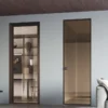 Two tall, modern ECLISSE 40 Hinged Aluminium Framed Glass doors with dark frames are set in a light wall. The left door opens to a room with shelves and decor, while the right reflects the space. A grey sofa is partially visible on the right.