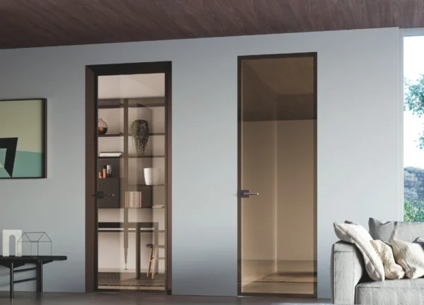 Two tall, modern ECLISSE 40 Hinged Aluminium Framed Glass doors with dark frames are set in a light wall. The left door opens to a room with shelves and decor, while the right reflects the space. A grey sofa is partially visible on the right.