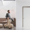 A man walks past a modern minimalist space with a wooden bench, books, magazines, two beige pouffes, and three pendant lights above. On the right, an ECLISSE 40 Timber pocket door is closed.
