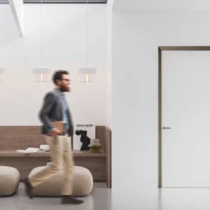 A man walks past a modern minimalist space with a wooden bench, books, magazines, two beige pouffes, and three pendant lights above. On the right, an ECLISSE 40 Timber pocket door is closed.