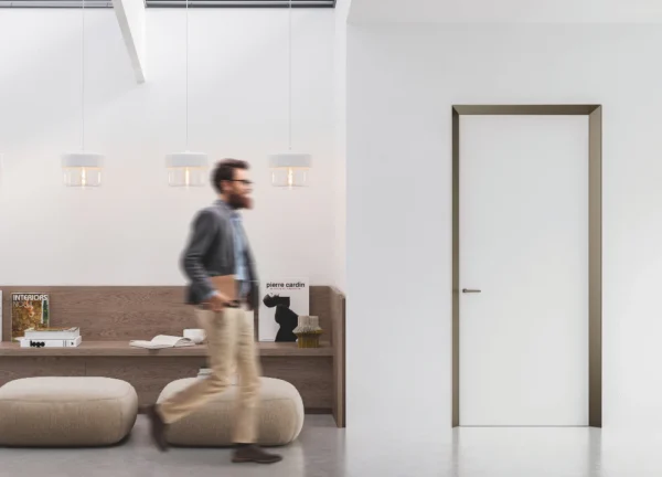 A man walks past a modern minimalist space with a wooden bench, books, magazines, two beige pouffes, and three pendant lights above. On the right, an ECLISSE 40 Timber pocket door is closed.