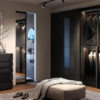 Modern walk-in wardrobe with glass wardrobe doors, neatly hung clothes, a sleek grey chest of drawers, ottoman and blanket, shoes on the floor, and an ECLISSE VetroLuxe Aluminium Framed Glass Pocket Door leading to a bathroom with a round mirror.