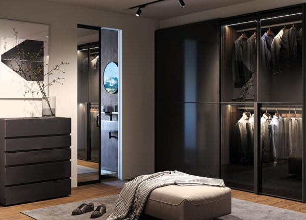 Modern walk-in wardrobe with glass wardrobe doors, neatly hung clothes, a sleek grey chest of drawers, ottoman and blanket, shoes on the floor, and an ECLISSE VetroLuxe Aluminium Framed Glass Pocket Door leading to a bathroom with a round mirror.