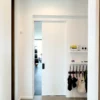 A bright corridor with white walls leads to a room through a partially open Cavity Slider Pocket Door Frame Kit. On the right, hooks with hanging bags and a small shelf with various items are mounted above them.