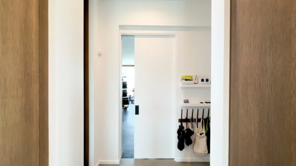 A bright corridor with white walls leads to a room through a partially open Cavity Slider Pocket Door Frame Kit. On the right, hooks with hanging bags and a small shelf with various items are mounted above them.