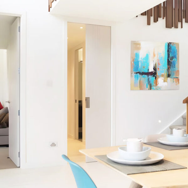 A modern dining area with a light wood table for two, an abstract painting, and a partially open cavity slider pocket door frame kit leading to the hallway. Neutral tones and clean lines give the space a minimalist feel.
