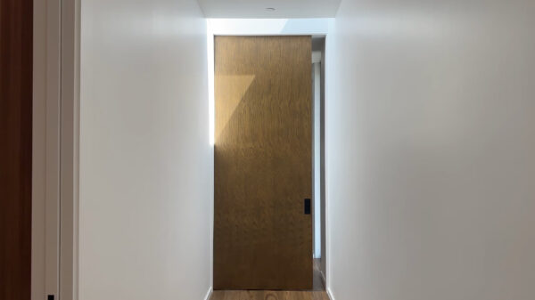 A narrow corridor with white walls and a wooden floor leads to a large sliding door fitted with the Cavity Slider Pocket Door Frame Kit, its minimalist, modern design partially illuminated by sunlight from above.